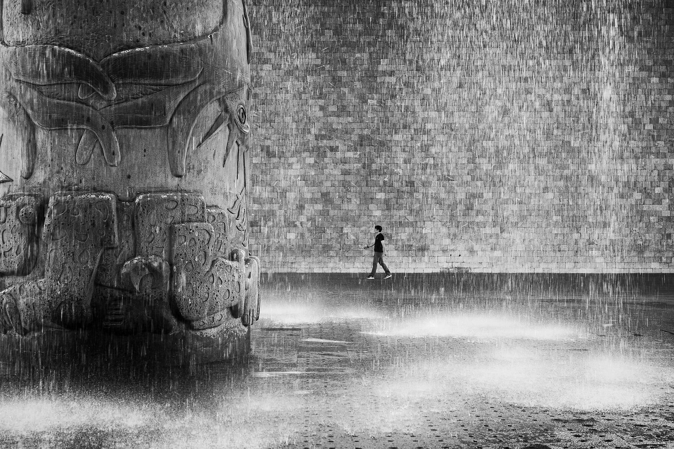 A lone figure walks beside a monumental carved stone as water falls from the museum’s vast concrete roof.
